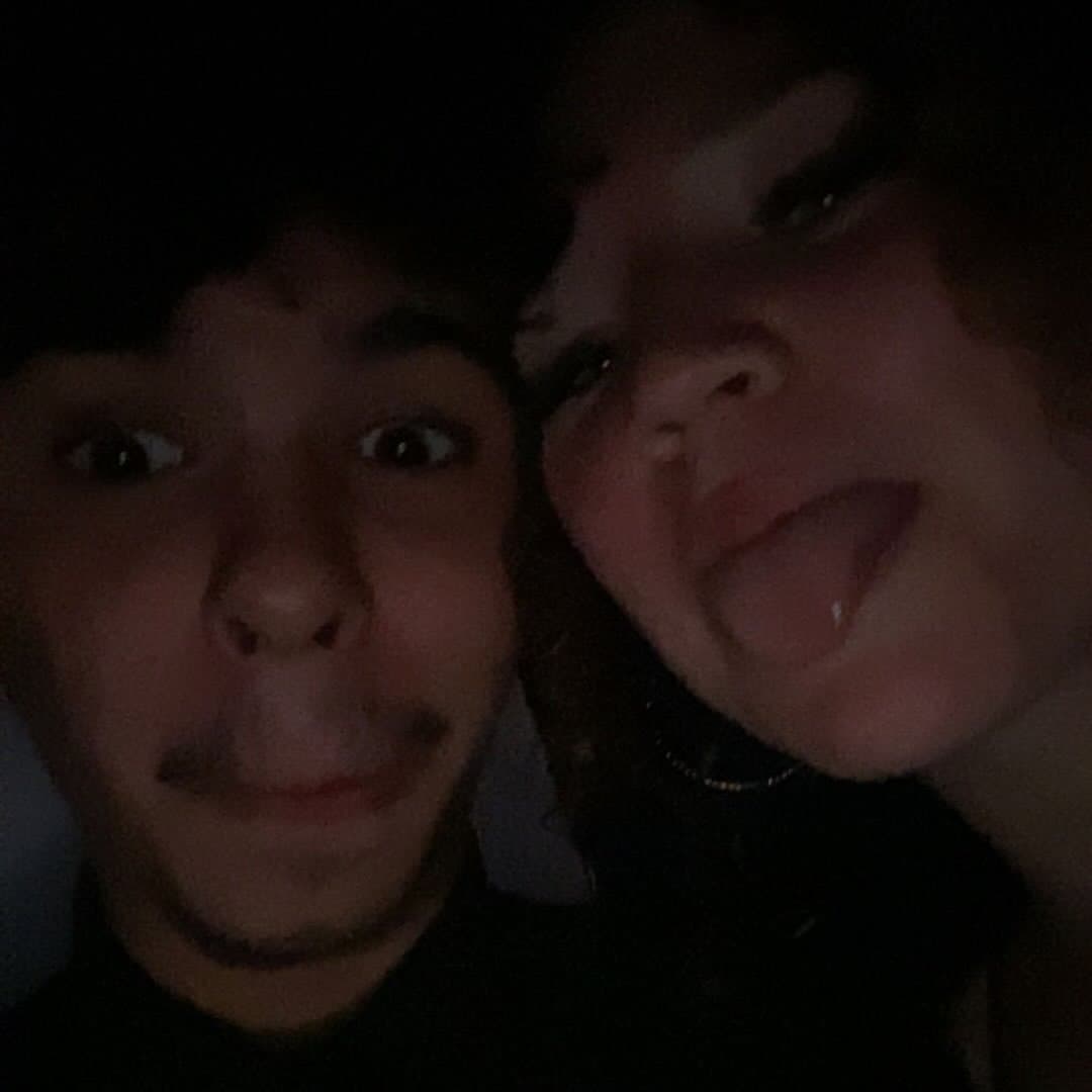 Low-light, close-up selfie of two young people, with one sticking her tongue out playfully.