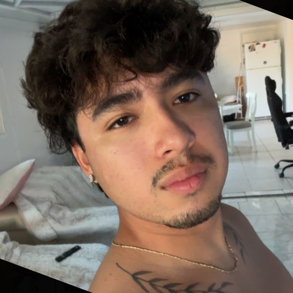 Young man with curly dark hair, goatee, and chest tattoo wearing a gold chain.