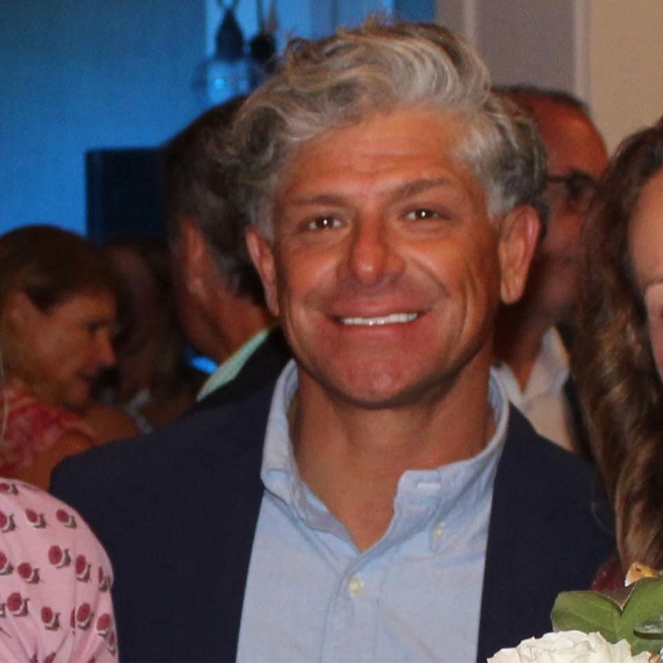 Smiling man with wavy grey hair wearing a navy blazer and a light blue shirt.